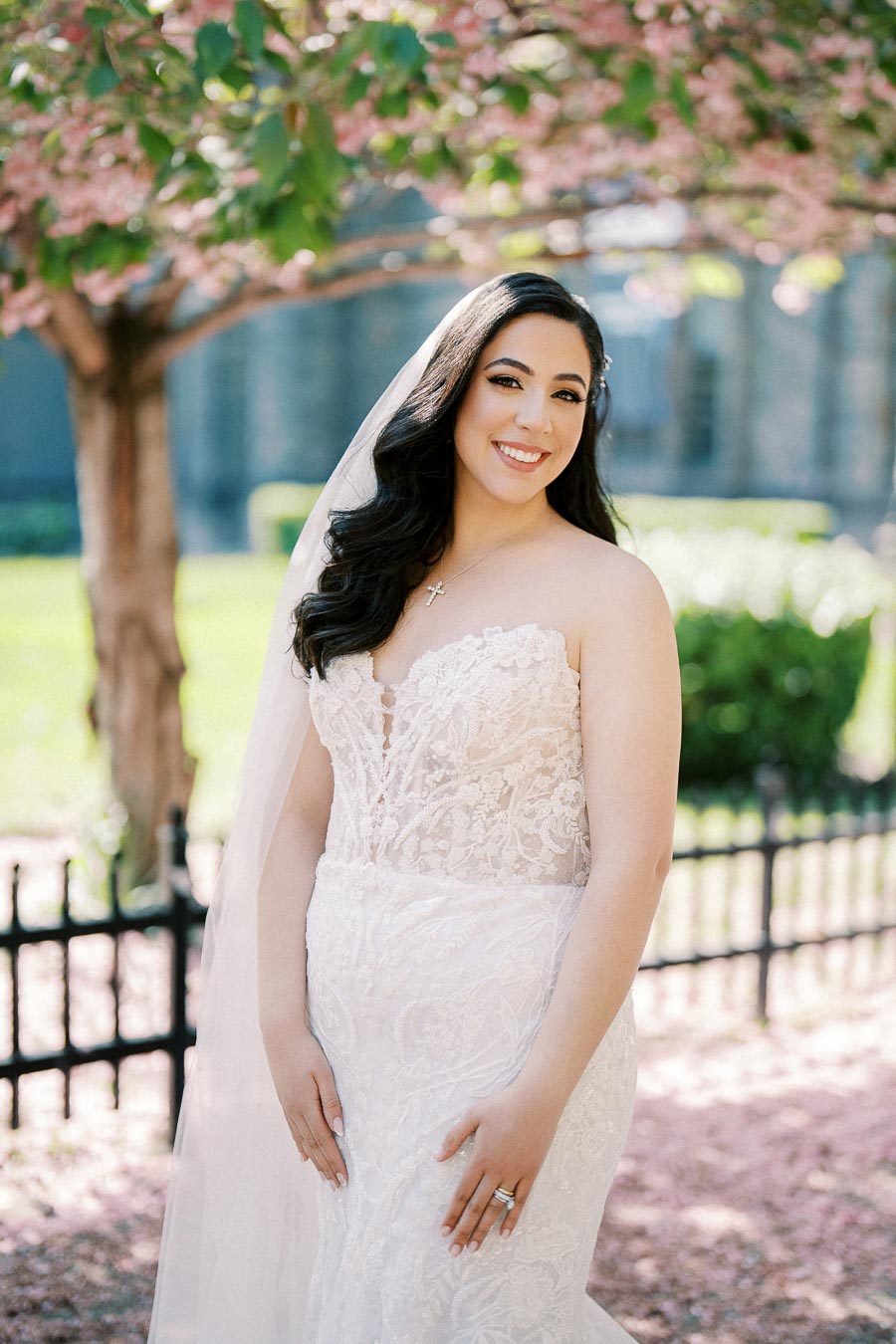 A bride in a lace wedding dress and veil poses outdoors in front of a blooming cherry blossom tree, with a soft smile.