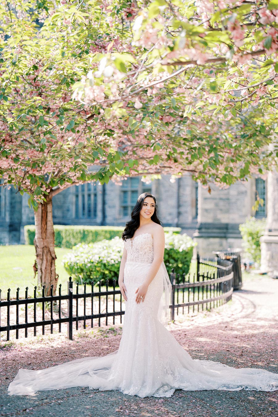 Bride in a white wedding dress standing under cherry blossom trees in a garden setting, with a wrought iron fence and historic stone building in the background.