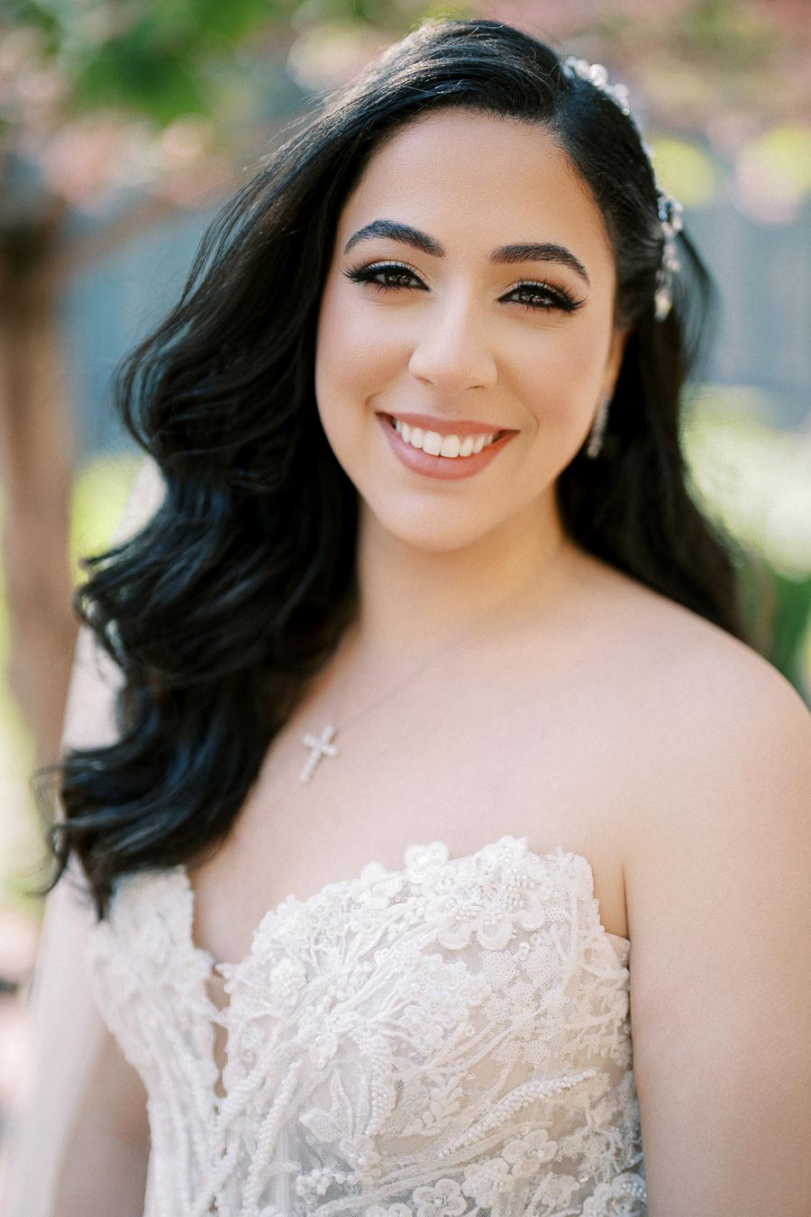 A smiling bride in a detailed lace wedding dress stands outdoors with softly blurred greenery in the background, showcasing elegant makeup and long dark hair styled in loose waves.