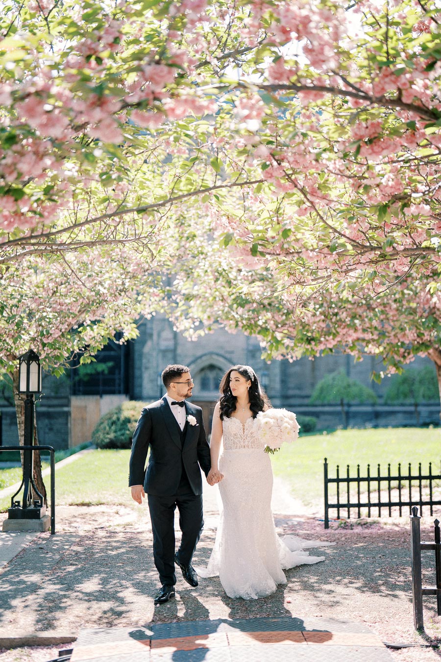 A newlywed couple walks hand in hand under blooming cherry blossom trees in a sunny garden, with the bride in a lace gown holding a bouquet and the groom in a dark suit.