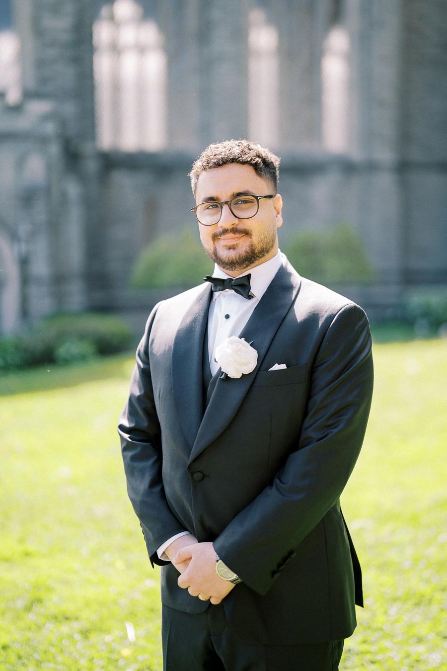 A man in a black tuxedo and bow tie stands smiling in a sunlit garden setting with a historic building in the background.