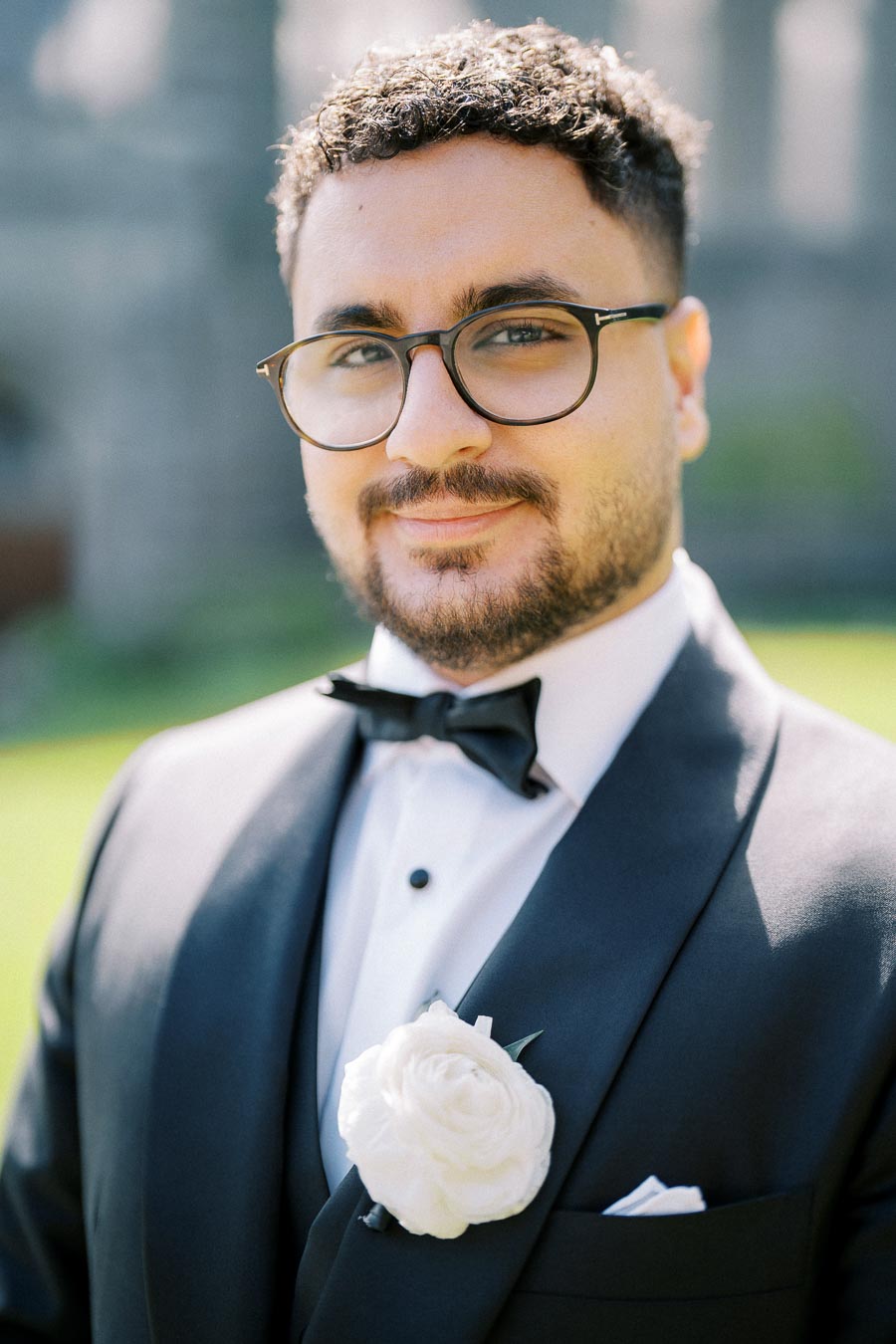 A man in a formal black suit with a white bow tie and a white rose boutonniere, smiling outdoors against a blurred green background.