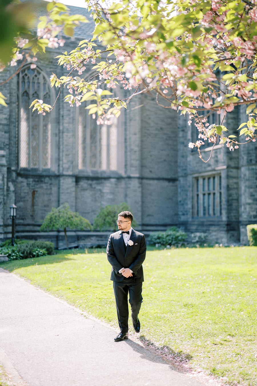 A groom in a black tuxedo walking along a path near a historic stone building, under a blooming cherry blossom tree on a sunny day.
