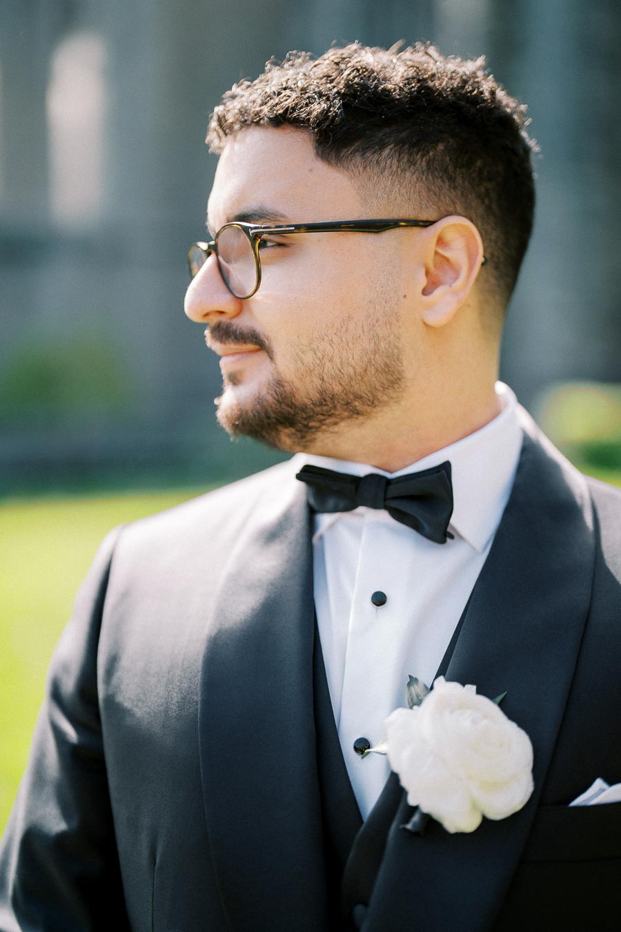 A groom in a black tuxedo with glasses and a rose boutonniere, looking to the side at an outdoor wedding.