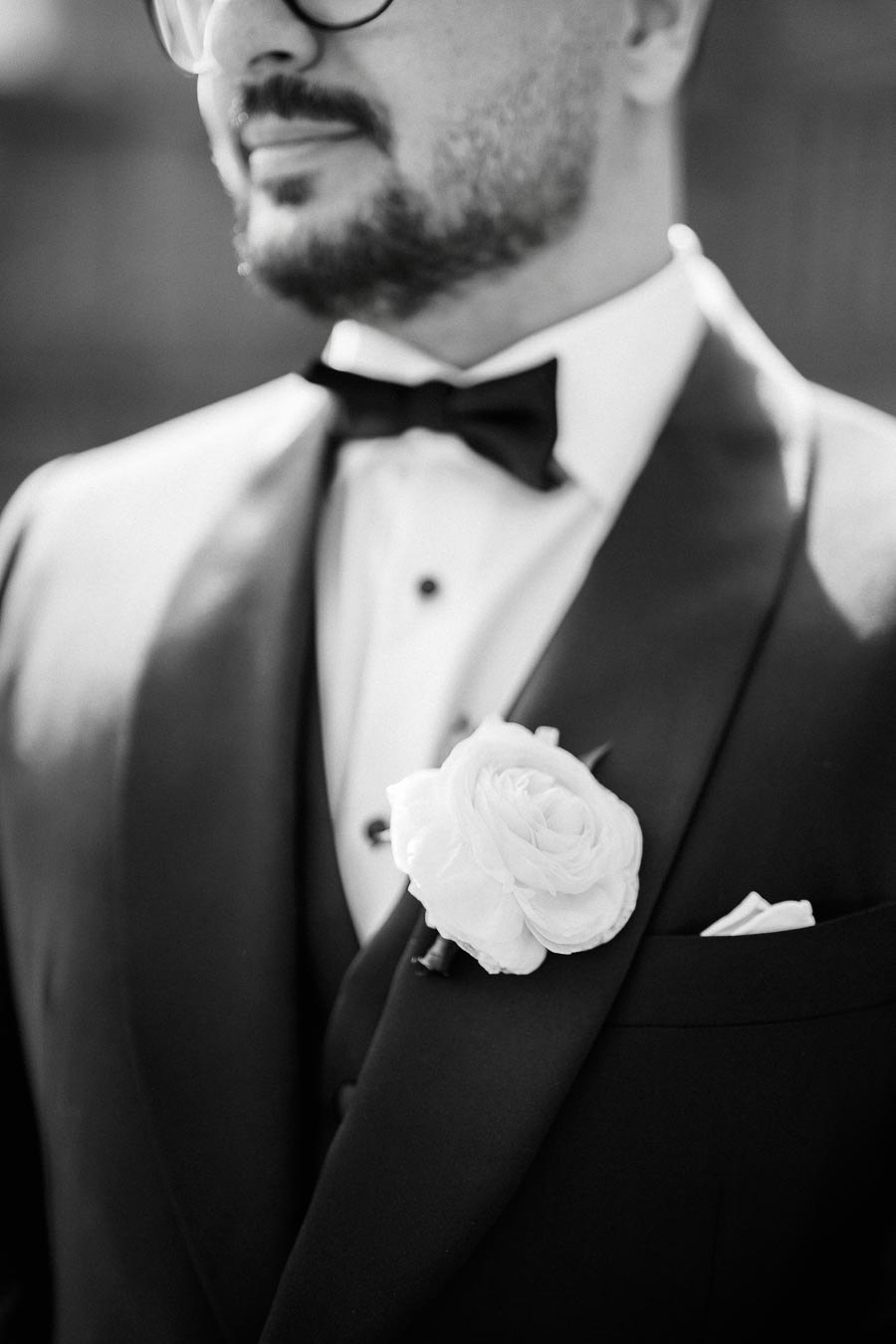 Black and white close-up of a groom in a tuxedo, featuring a white rose boutonniere and a bow tie.