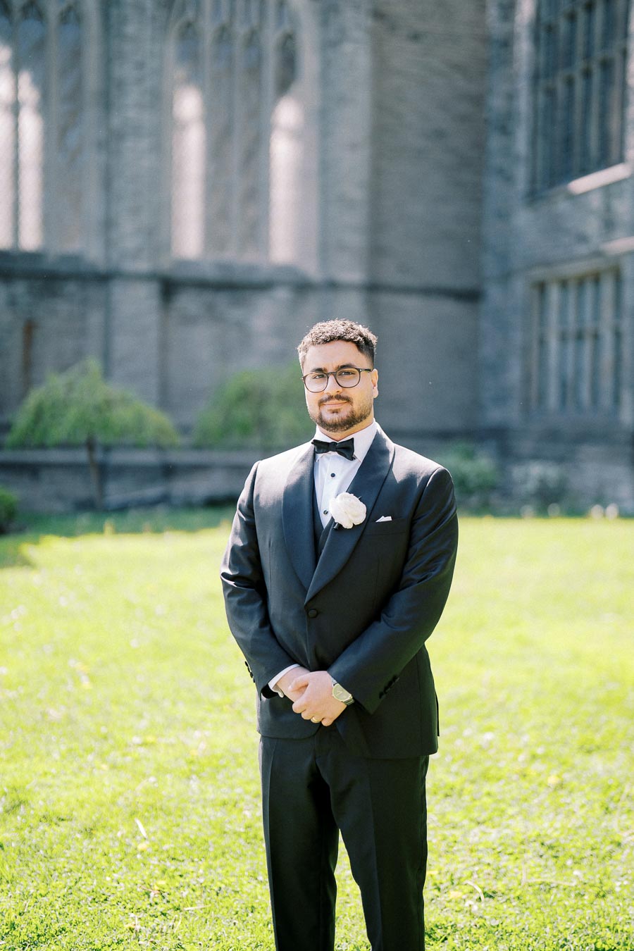 A man in a black tuxedo with a white boutonniere stands on a lawn in front of a stone building, possibly a church or cathedral, on a sunny day.