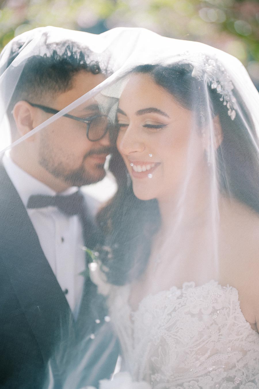 Romantic wedding couple embracing under a veil, bride in lace gown with jeweled details, smiling and groom in a tuxedo, capturing an intimate moment.