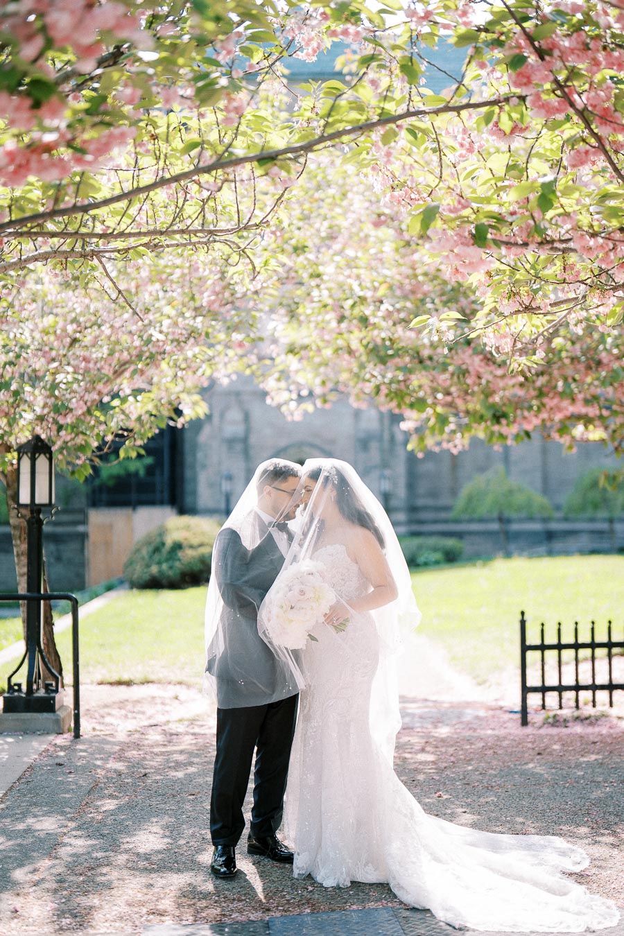 Romantic wedding couple embraces under floral archway with sunlight filtering through cherry blossoms, bride in elegant lace gown holding white bouquet.