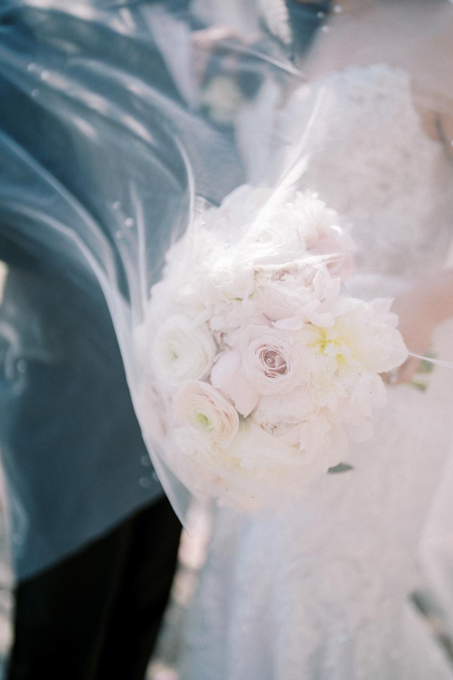Bridal couple embracing with a focus on a delicate bouquet of white and pastel pink roses, enveloped in a soft, flowing veil.