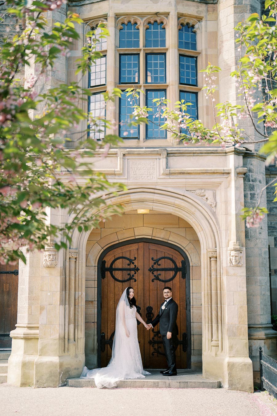 Elegant wedding couple standing in front of a historic stone building with ornate wooden doors, surrounded by blossoming trees.
