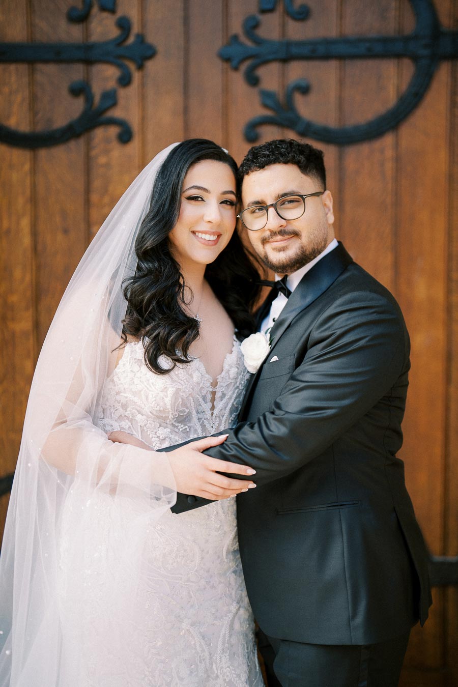 A newlywed couple embracing in front of a rustic wooden door, with the bride wearing an elegant lace wedding dress and veil, and the groom in a classic black suit and tie, both smiling warmly.