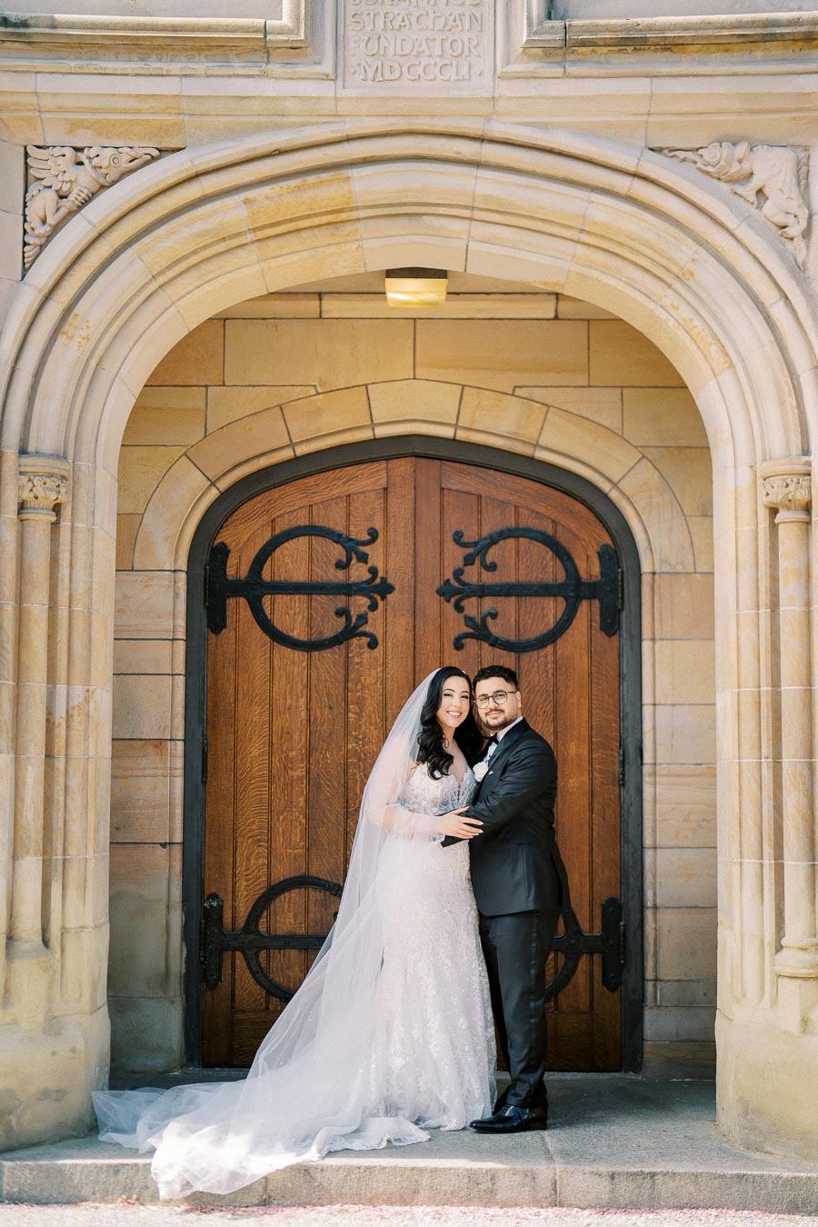 A newlywed couple standing in front of an ornate wooden church door, surrounded by detailed stone architecture. The bride wears a flowing white gown and veil, while the groom is dressed in a classic black suit.