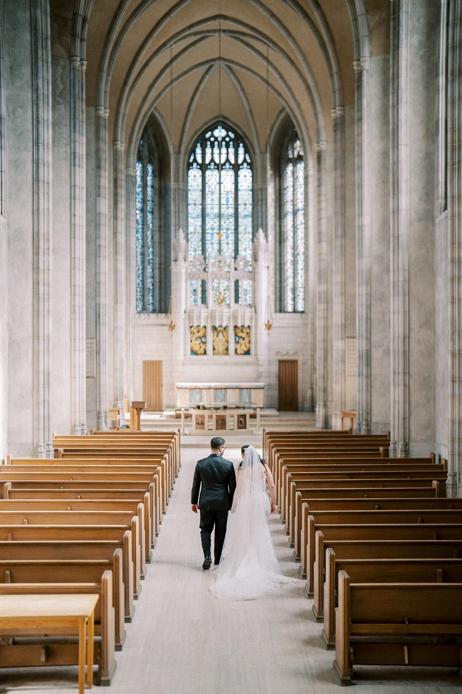 A bride and groom walking down the aisle of a grand cathedral with high arches and stained glass windows.