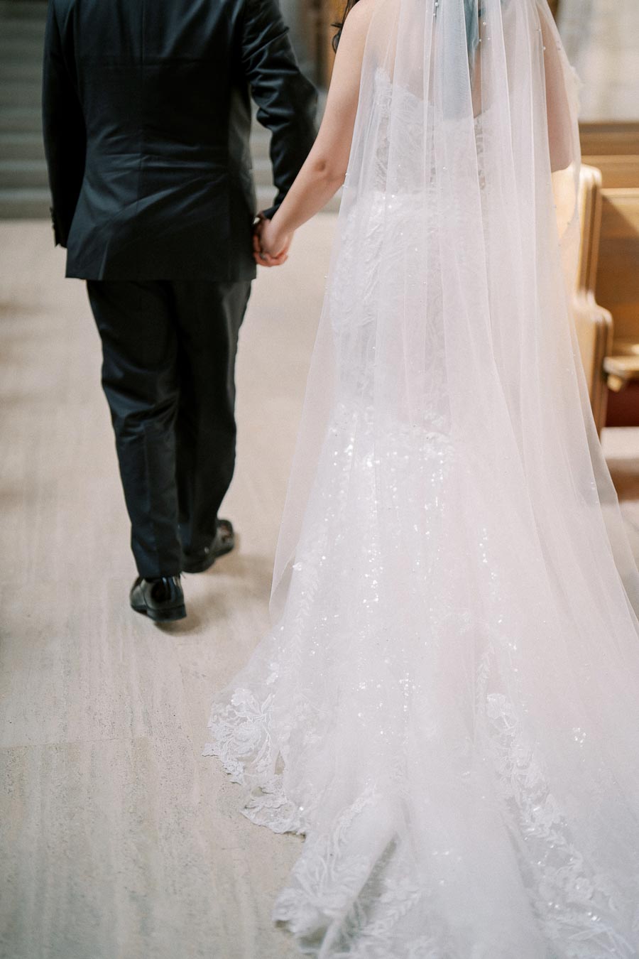 Bride and groom walking hand in hand down a church aisle, showcasing the bride's elegant white gown and lace veil trailing behind.