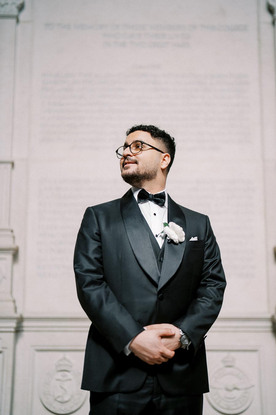 A groom in a black tuxedo with a white boutonniere stands confidently in front of an intricately designed wall, looking to the side with a slight smile.