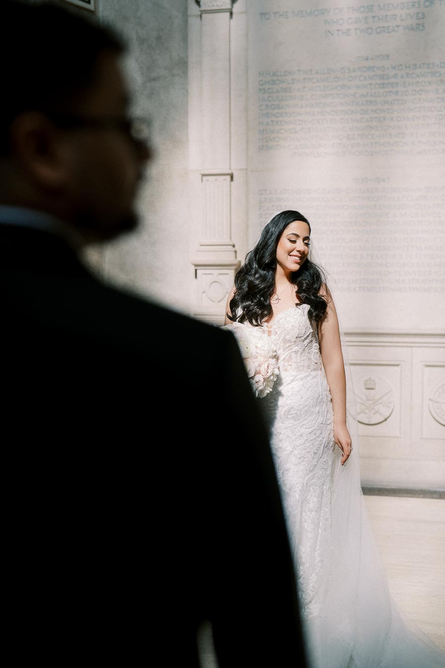 Bride in an elegant white lace gown smiling gracefully under soft natural light, with a blurred figure in the foreground, standing in front of a stone memorial wall.
