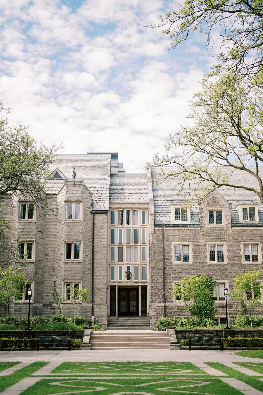 Alt text: Historic stone building with large windows and slate roof surrounded by greenery and trees under a partly cloudy sky.
