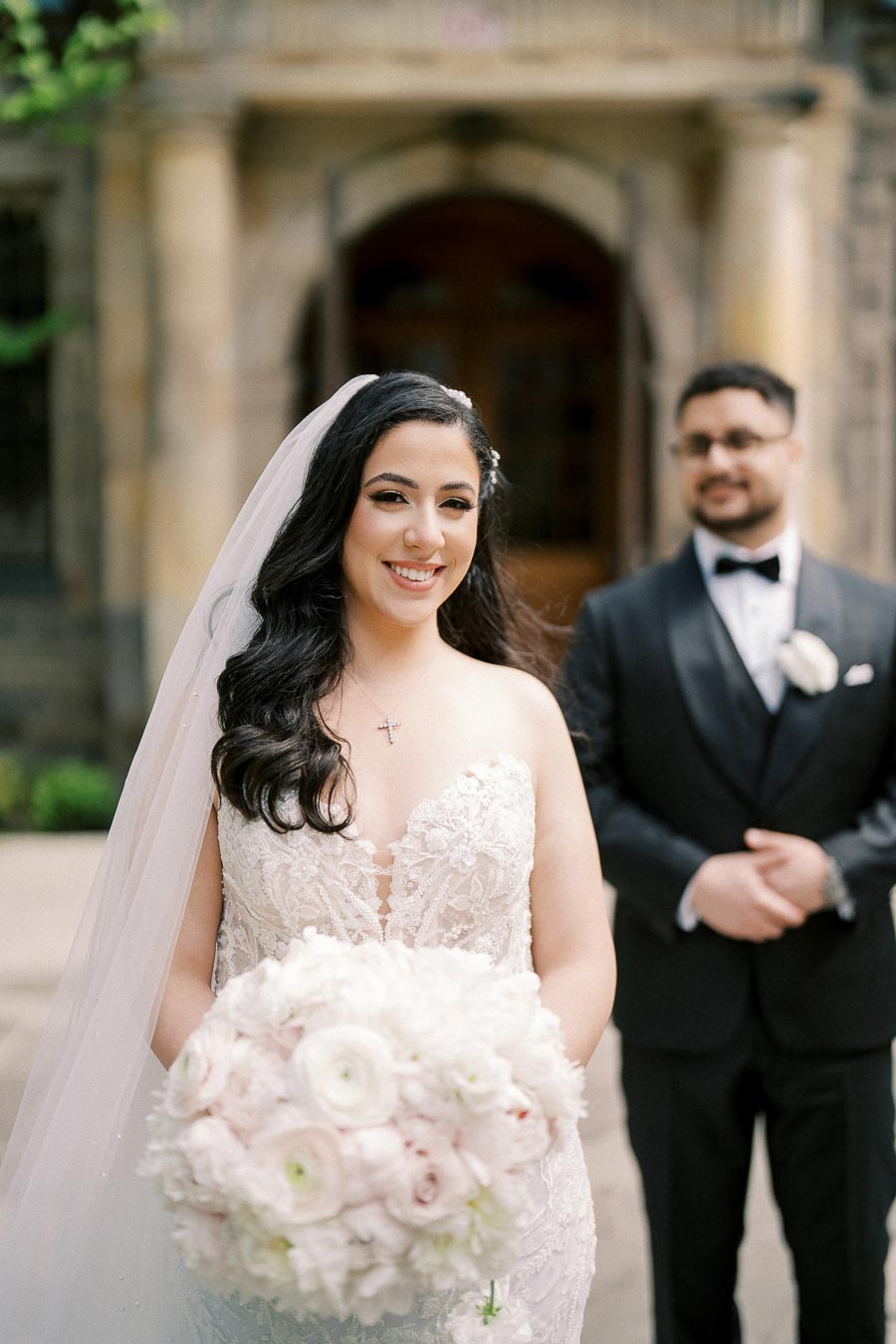 Bride in elegant lace wedding gown smiling while groom stands in background wearing a black suit and bow tie, holding a bouquet of white and pink flowers in front of a historic building.