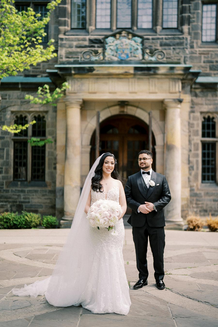 Bride and groom posing in front of an elegant stone building with greenery, the bride holding a bouquet of white flowers, and both dressed in formal wedding attire.
