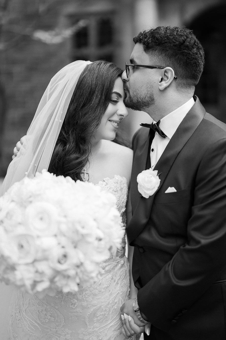 A black and white wedding photo of a couple lovingly embracing. The bride is in a lace wedding dress with a veil, holding a bouquet of flowers, while the groom wears a tuxedo and gently kisses her forehead.