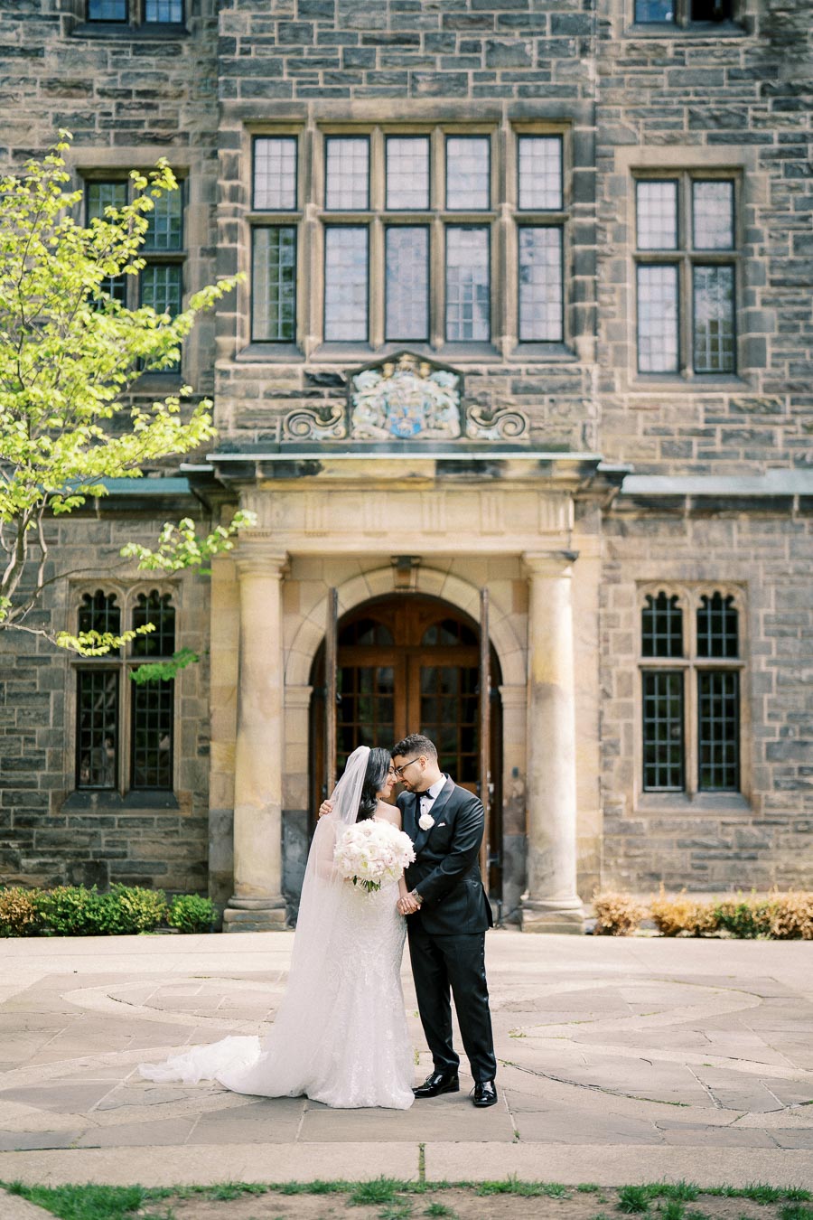 A bride and groom share an intimate moment in front of a historic stone building with large windows. The bride wears a white dress with a veil and holds a bouquet, while the groom is dressed in a black tuxedo. Lush greenery frames the scene, capturing a romantic wedding day atmosphere.