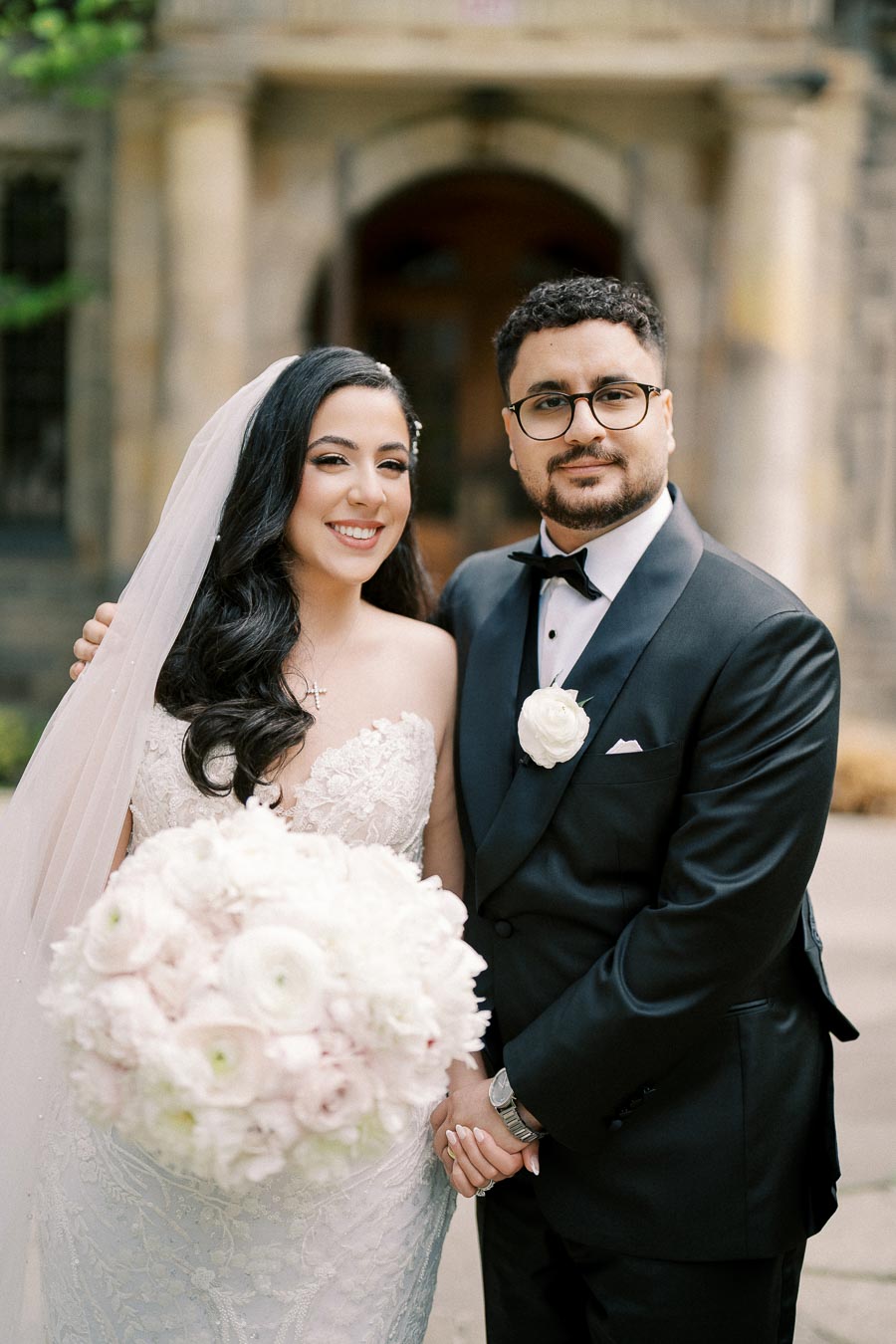 Elegant bride and groom posing on their wedding day, with the bride in a stunning lace dress holding a large bouquet of pink and white flowers, and the groom in a classic black tuxedo.