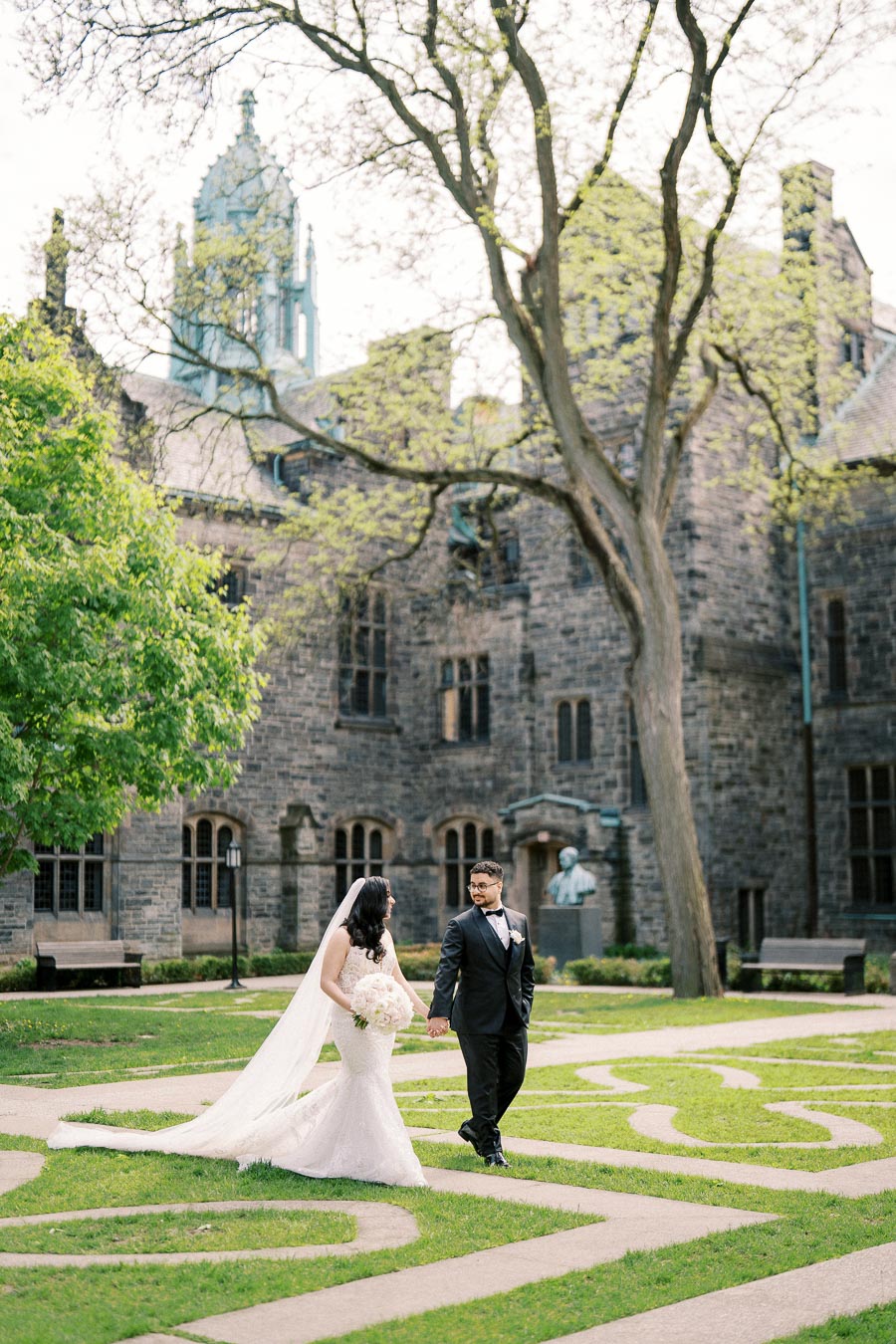 Elegant bride and groom walking hand in hand in a picturesque garden setting with historic stone building in the background, capturing a romantic wedding moment.