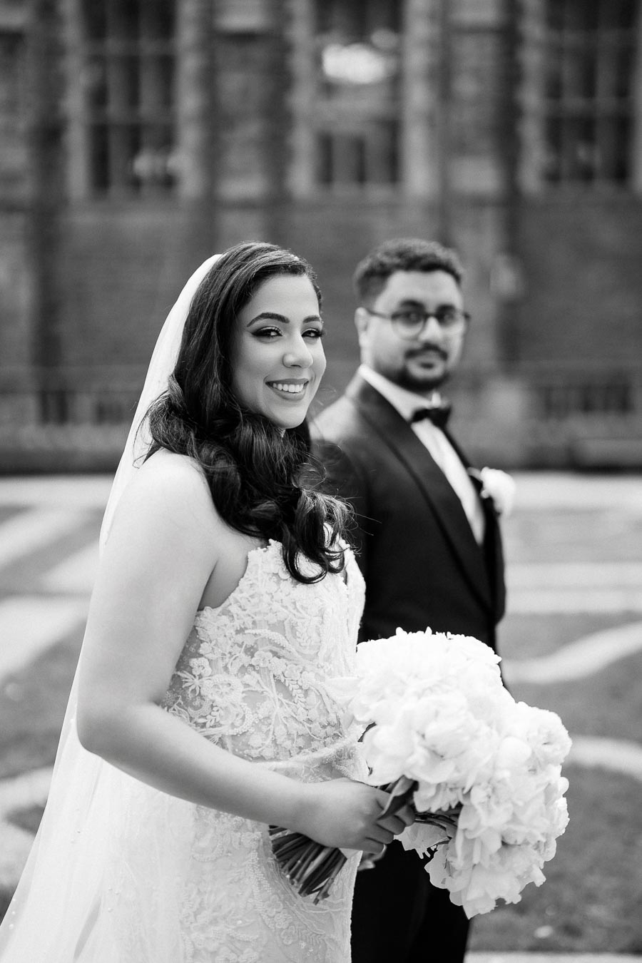 Black and white wedding photo of a smiling bride in a lace gown holding a bouquet, with a groom in a tuxedo in the background, standing outside near a historic building.