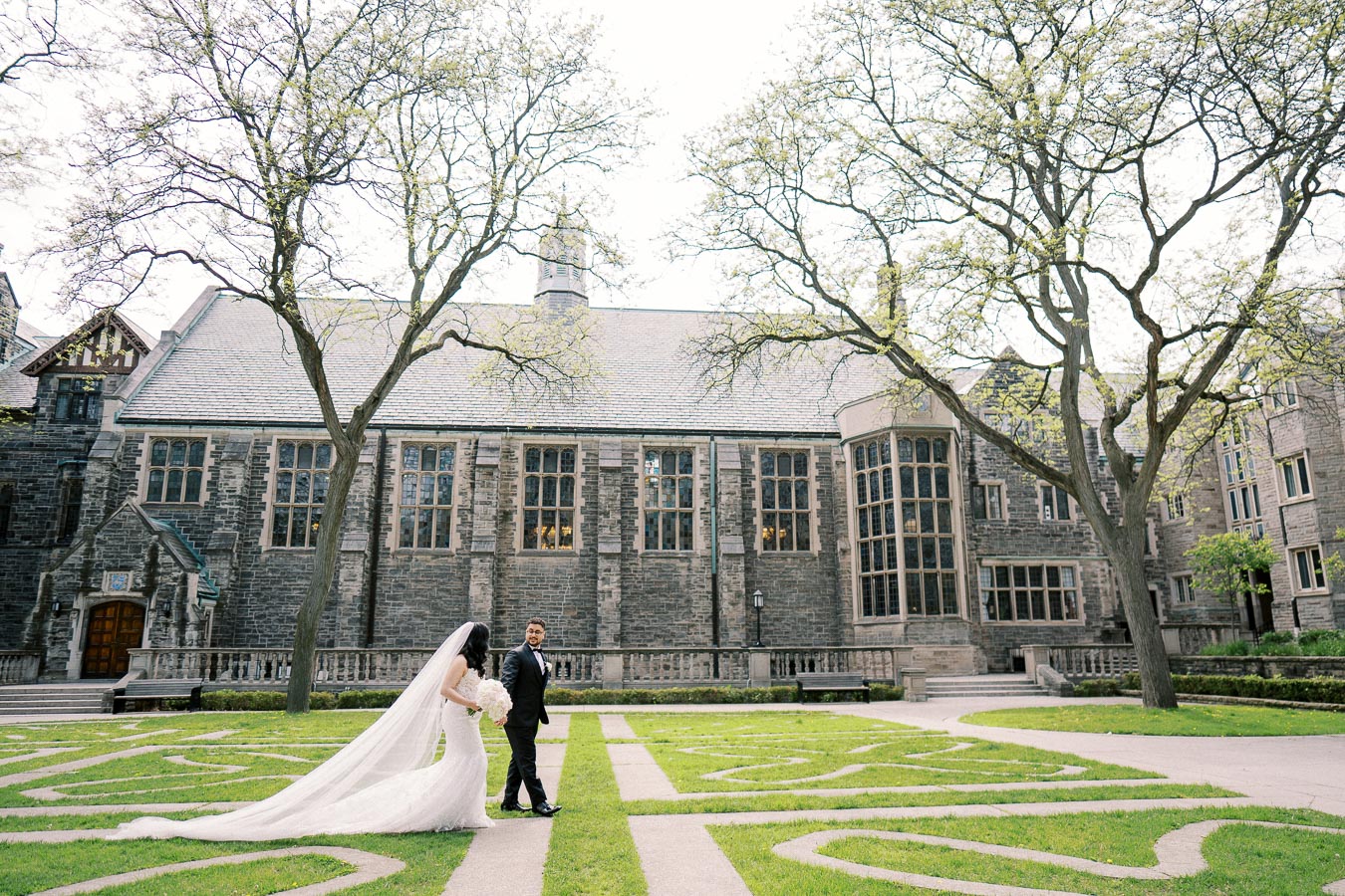Bride and groom holding hands in front of a historic stone building with large windows and a manicured garden, perfect for a wedding photoshoot.