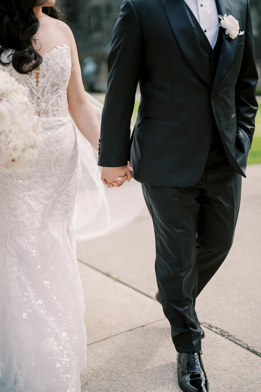 Elegant bride in a lace wedding gown holding hands with groom in a black tuxedo during outdoor ceremony.