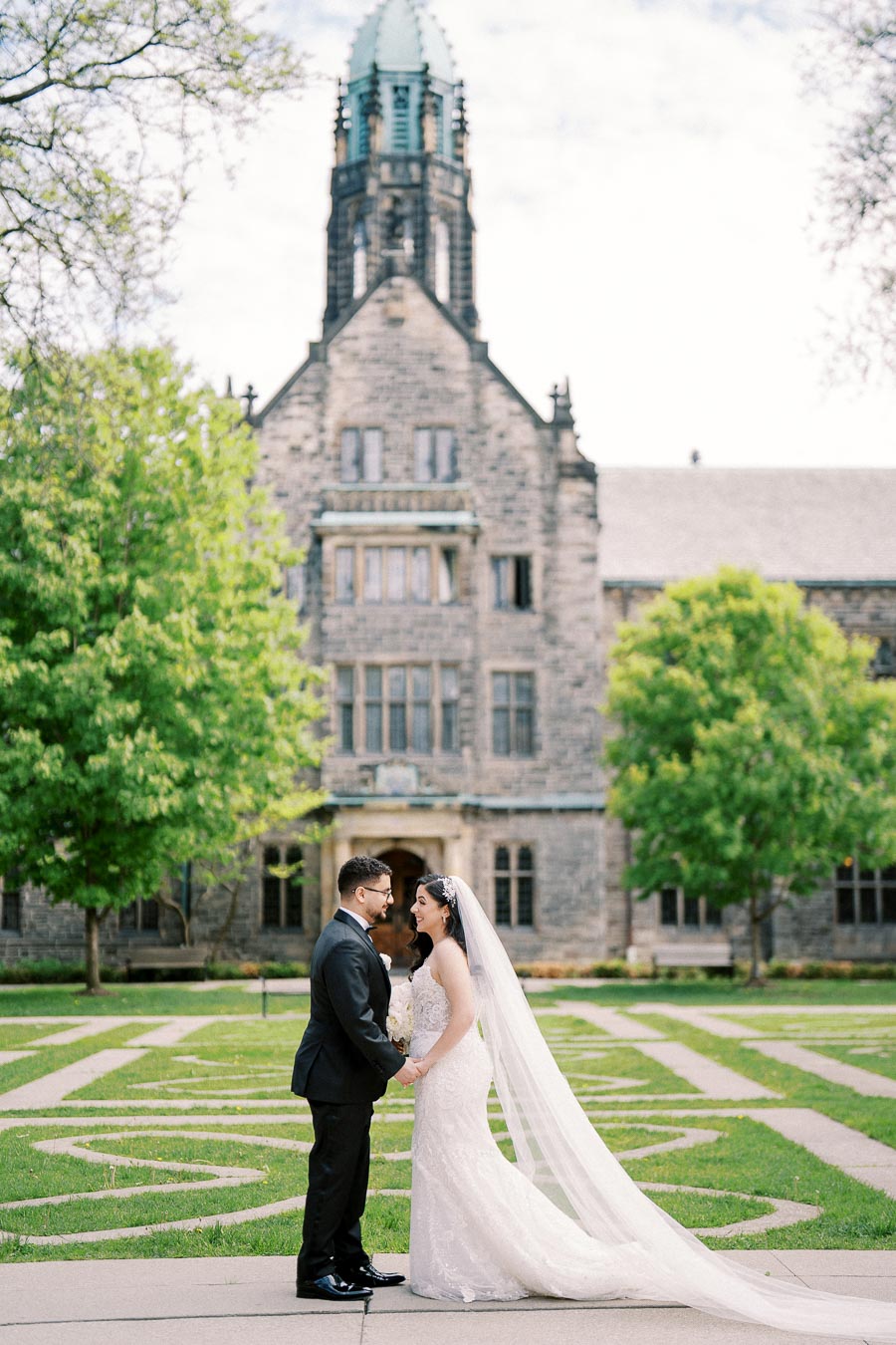 A bride and groom standing in front of a historic stone building with lush greenery, capturing a romantic wedding moment. The bride wears a long white gown with a veil, while the groom is in a black suit.