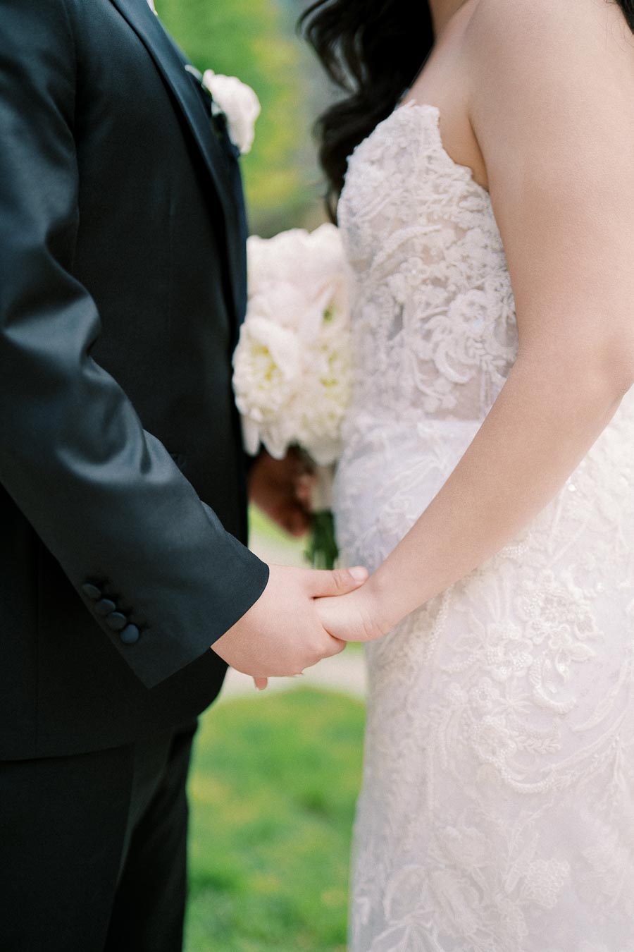 Close-up of a bride and groom holding hands, bride in a lace wedding dress and groom in a black suit, symbolizing unity and love on their wedding day.