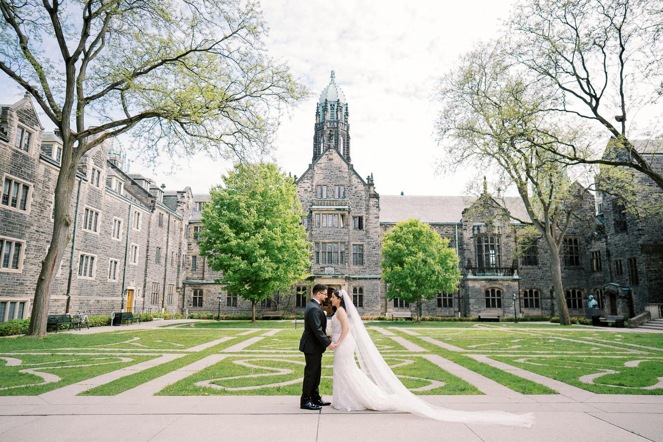 A bride and groom stand close together in front of a historic stone building, surrounded by green trees and grass on a clear day. The bride wears a long white dress with a flowing veil, and the groom is in a black suit. The scene captures a romantic wedding moment in a picturesque courtyard setting.