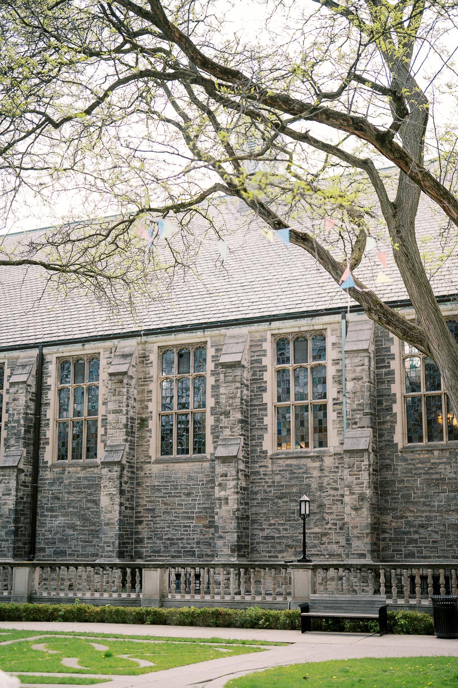 Alt text: Historic stone building with gothic-style windows, adjacent to a lush green lawn and a tree with budding leaves, under a clear sky.