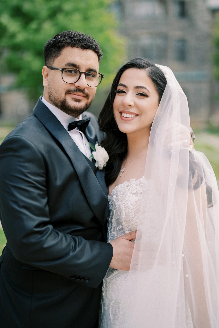 Smiling bride and groom in elegant wedding attire, posing outdoors with a blurred green background.