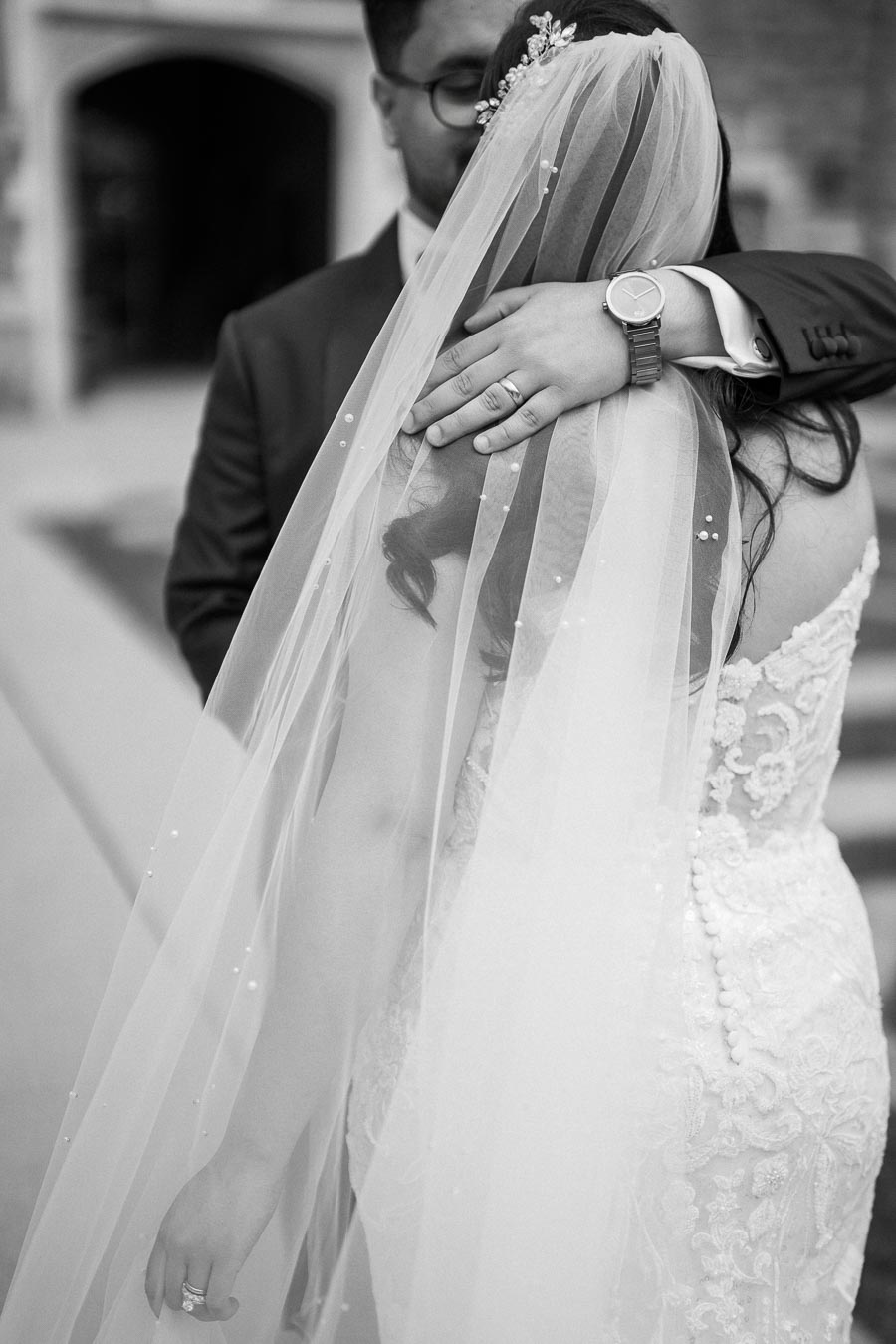 Black and white photo of a bride and groom embracing, focusing on the bride's detailed lace wedding dress and veil, with the groom's watch and wedding ring visible.