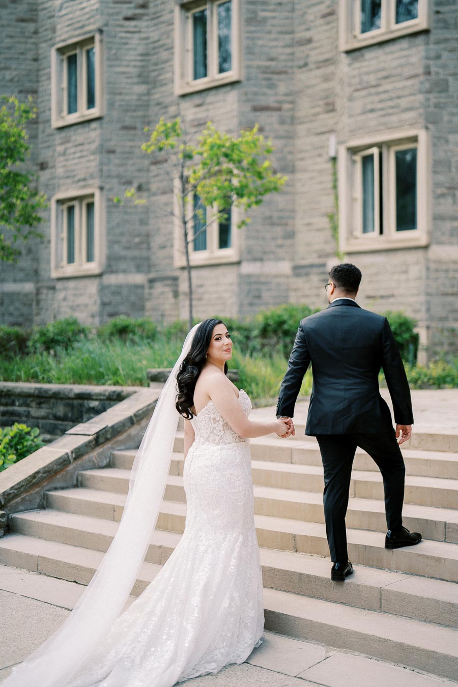 A bride in a lace wedding gown and long veil holds hands with a groom in a black suit as they ascend stone steps in front of a historic stone building, surrounded by greenery.