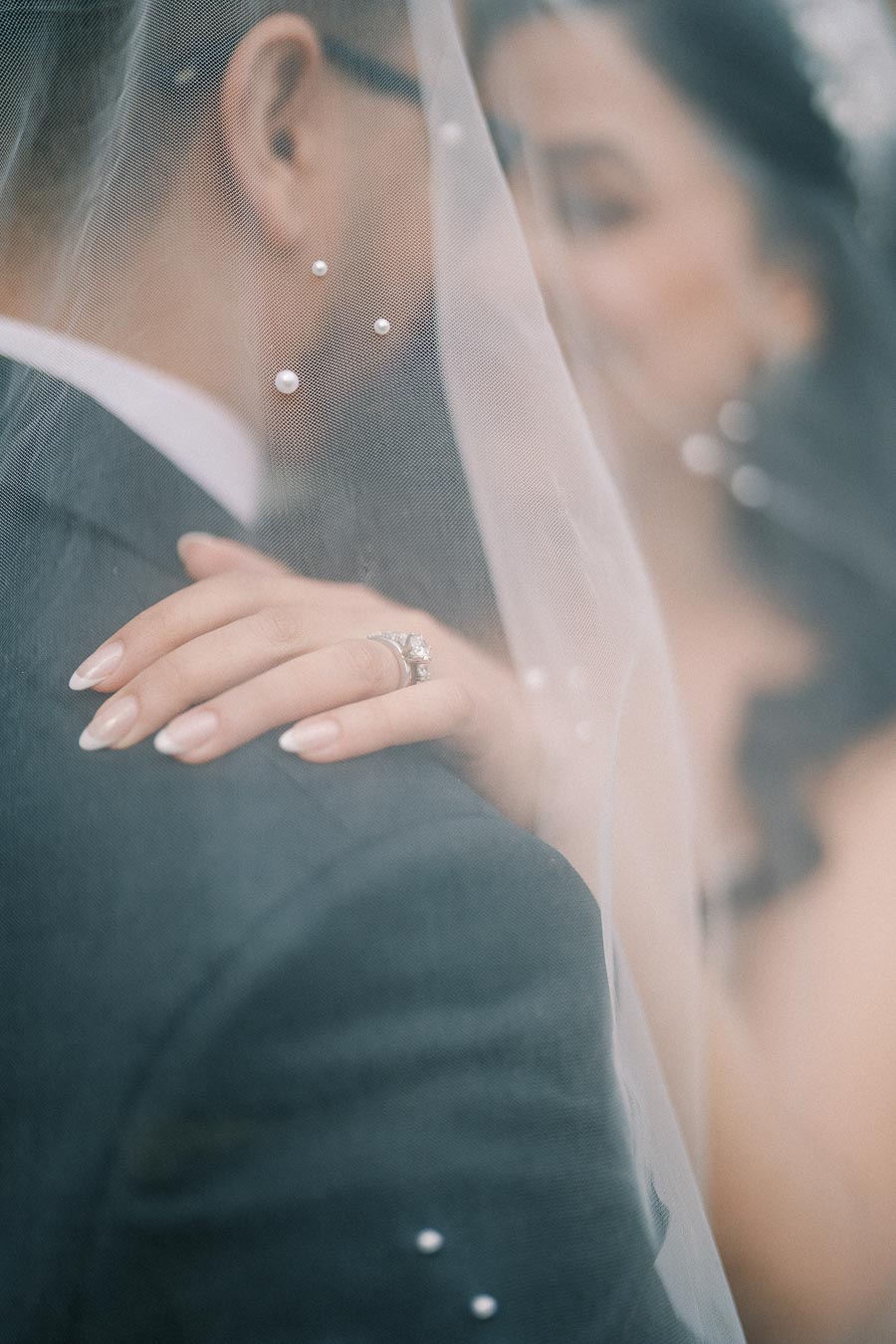 A bride and groom are softly embraced under a delicate wedding veil decorated with pearls. The bride's manicured hand rests on the groom's shoulder, showcasing an elegant engagement ring. The image captures an intimate and romantic moment.