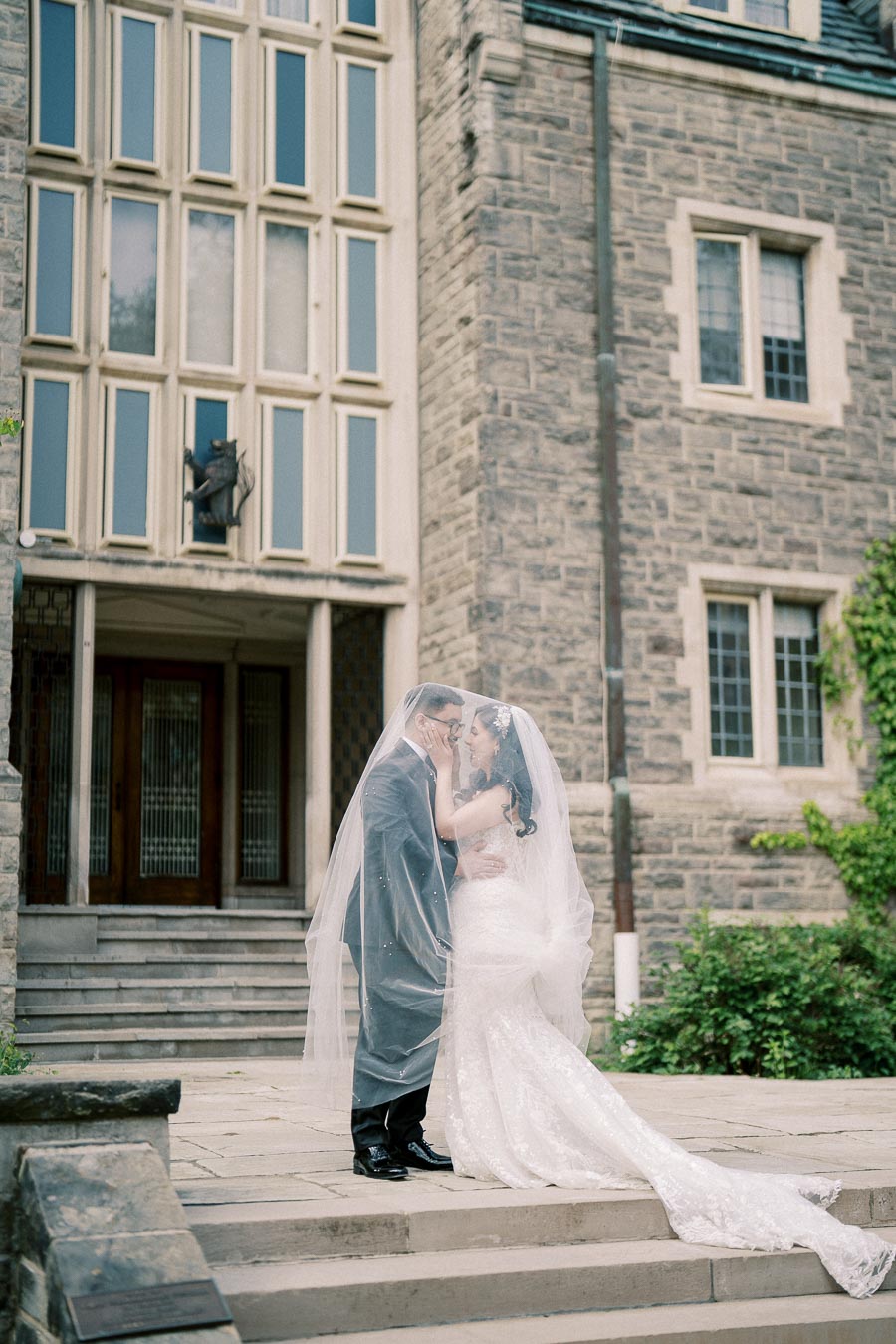 A bride and groom embrace under a delicate veil, standing on the stone steps of a historic building with large windows and ornate architecture, surrounded by lush greenery.