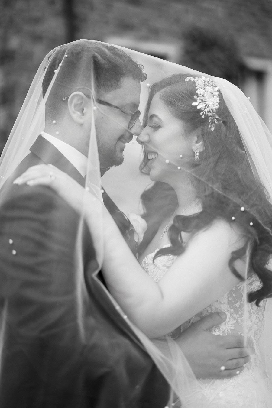 Romantic bride and groom embrace tenderly under a veil, showcasing love on their wedding day, with elegant bridal attire and floral hair accessory set against a soft background.