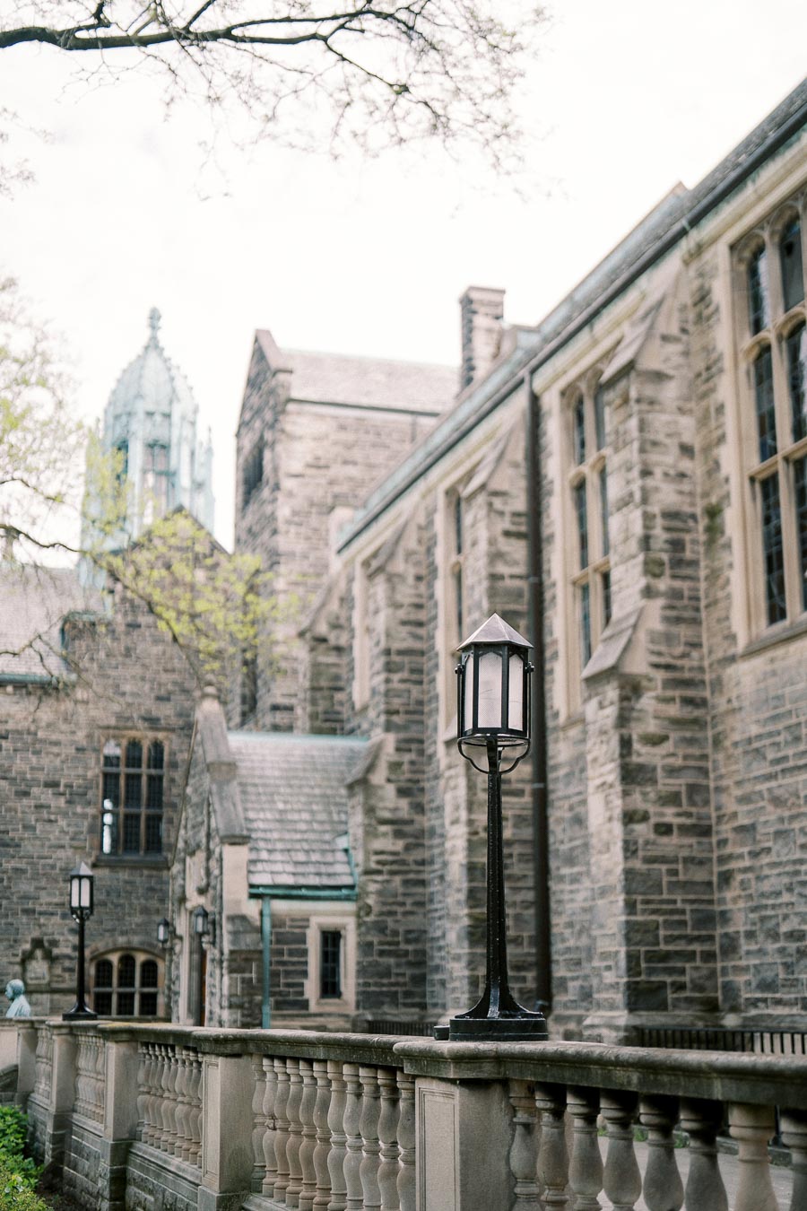 Gothic-style stone building with tall windows and a pointed roof, featuring a foreground of ornate street lamps and a decorative stone railing, under a soft, overcast sky.