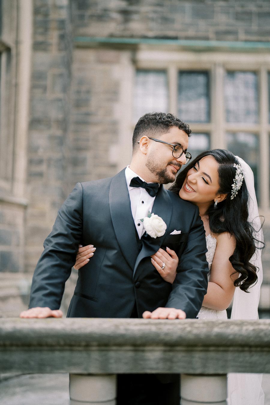 Bride and groom embracing joyfully in front of an elegant stone building, with the bride wearing a white gown and veil, and the groom in a black tuxedo.