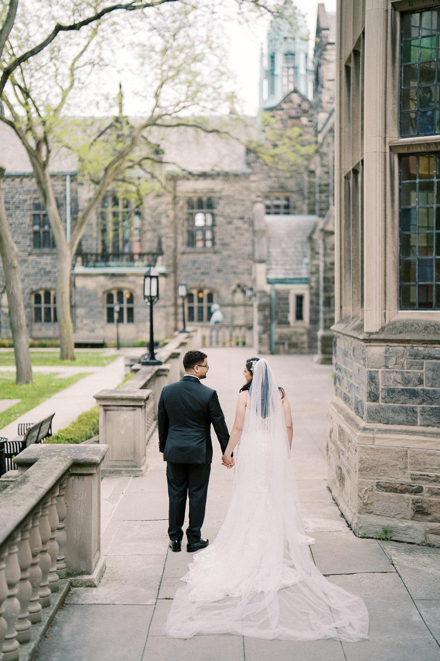 Bride and groom holding hands, walking in an elegant outdoor setting with stone architecture and greenery, perfect for wedding photography.