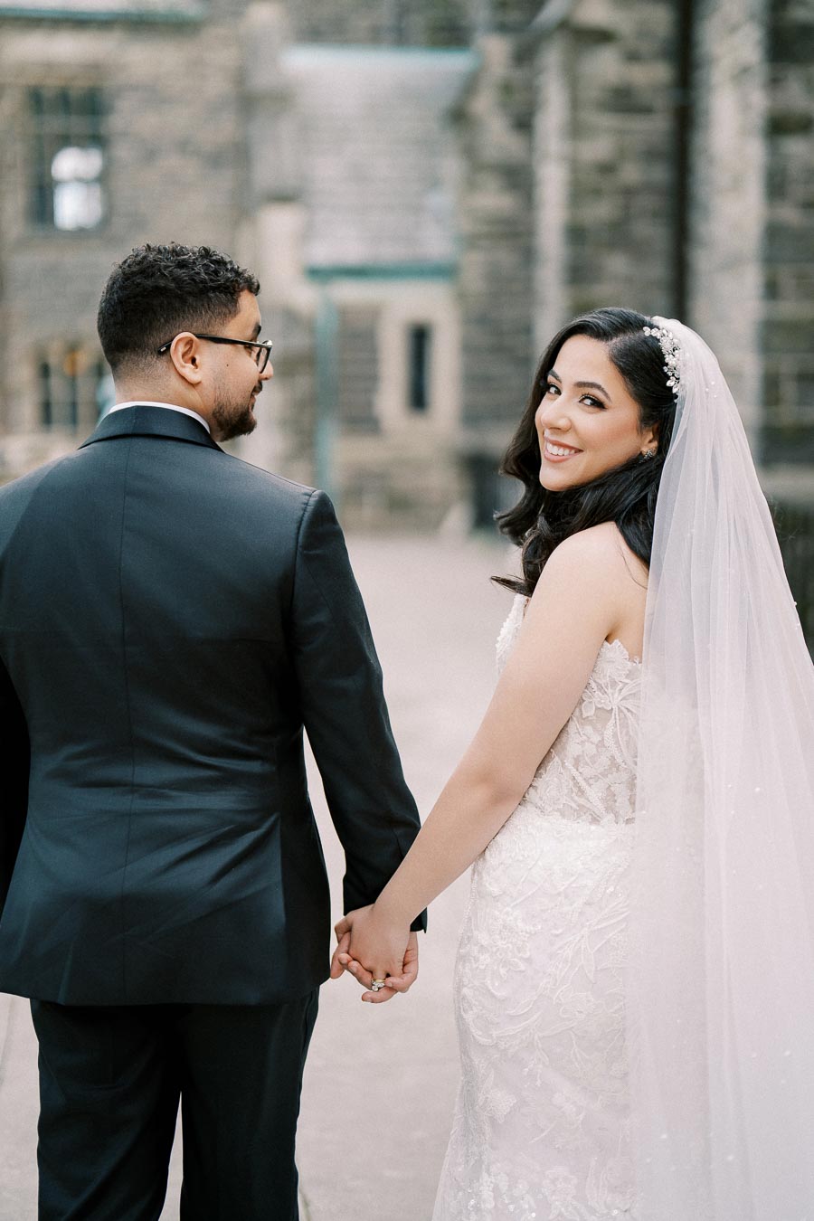 A bride and groom holding hands and smiling on their wedding day, standing in front of a stone building, with the bride wearing a lace dress and veil and the groom in a black suit.