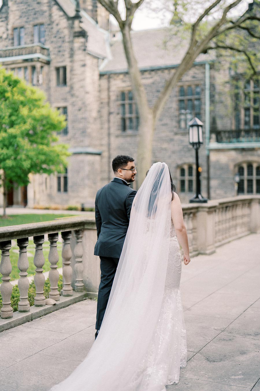 A bride and groom walking hand in hand along a stone balcony, with the bride in a flowing white gown and veil, and a historic stone building in the background.