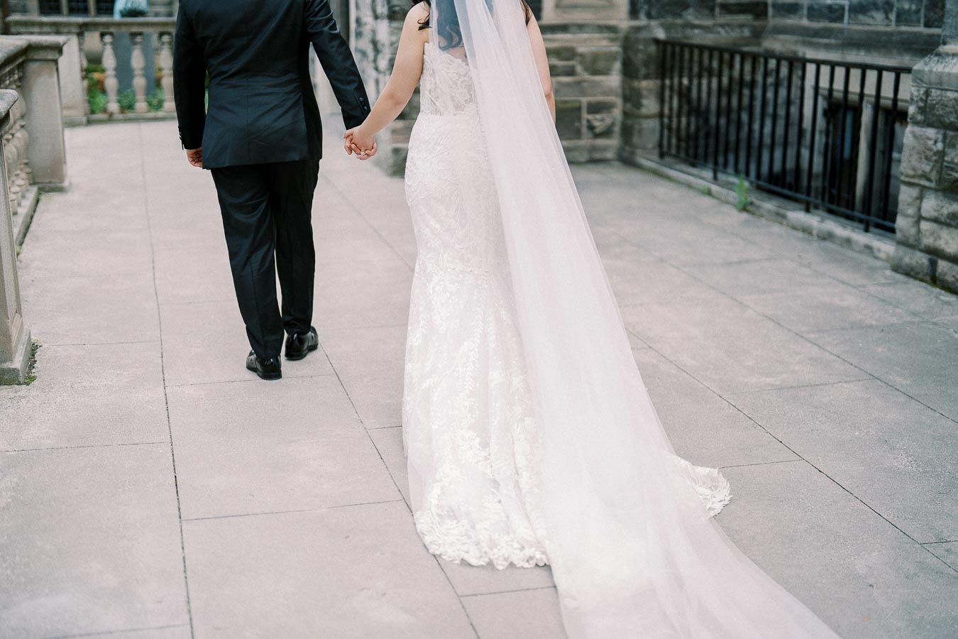Elegant bride and groom holding hands, walking together on a stone pathway, with the bride wearing a flowing white wedding dress and veil.