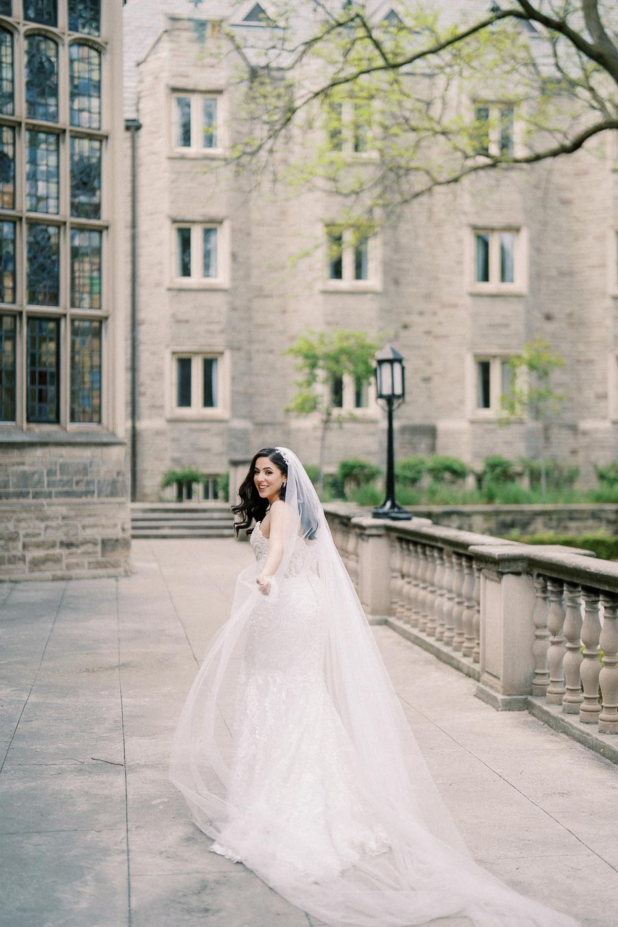 A bride in a flowing white gown and long veil smiles while walking outside a historic stone building, surrounded by lush greenery and ornate windows.