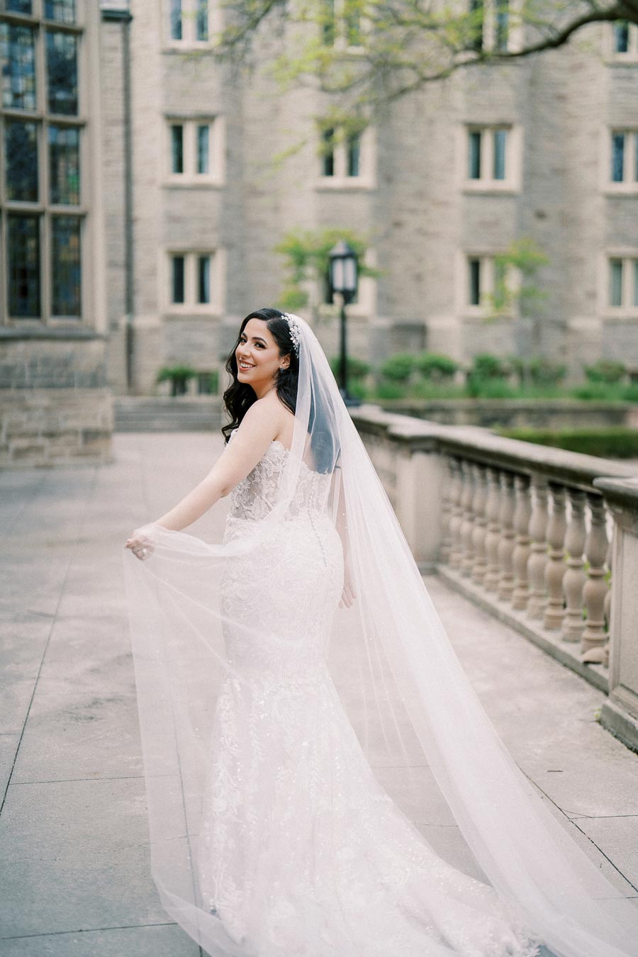 A bride in a lace wedding dress with a long veil smiles while walking outside an elegant stone building, surrounded by greenery.