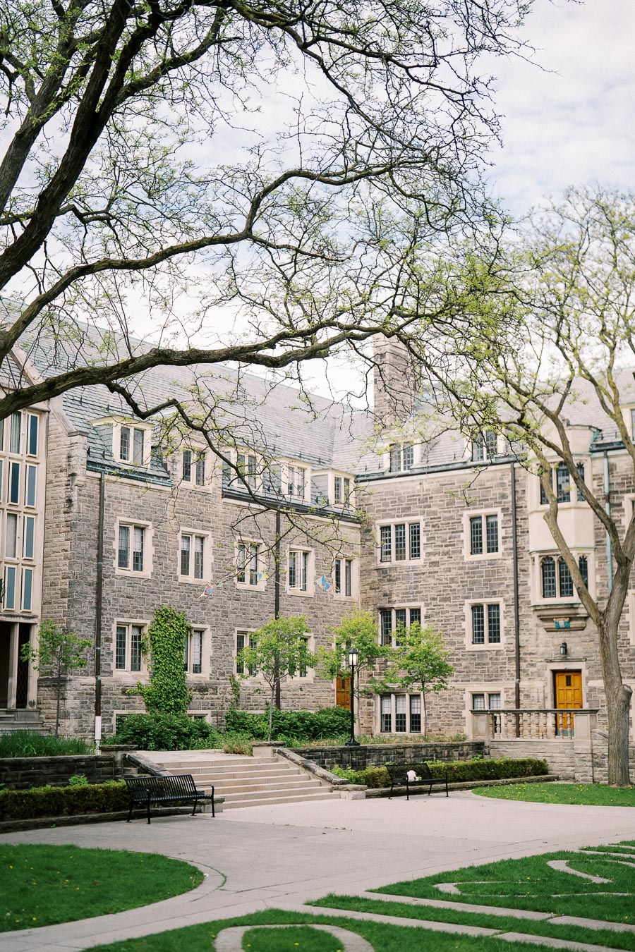 Historic stone building with arch windows, surrounded by green trees and a neatly landscaped courtyard.