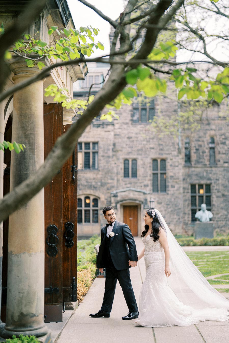 A newlywed couple in elegant wedding attire walks hand in hand outside a historic stone building, surrounded by green foliage and an enchanting garden.